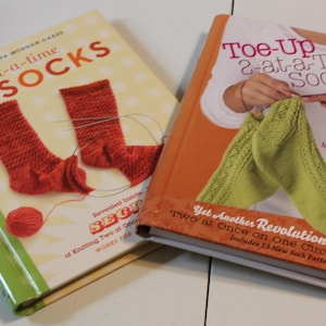 May include: Two books with the title "2-at-a-time Socks" on the cover. The book on the left is red and green and shows two red socks. The book on the right is orange and green and shows two green socks.