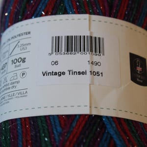 May include: A close-up of a ball of Vintage Tinsel yarn. The yarn is a mix of red, blue, and green with silver metallic threads. The label indicates it is 100g and has the text "Vintage Tinsel 1051".