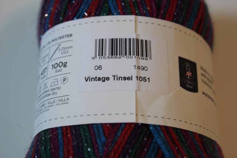 May include: A close-up of a ball of Vintage Tinsel yarn. The yarn is a mix of red, blue, and green with silver metallic threads. The label indicates it is 100g and has the text "Vintage Tinsel 1051".