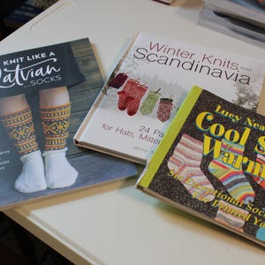 May include: Three knitting pattern books with colorful covers. The first book is titled "Knit Like a Latvian...Socks" and features a pair of colorful socks. The second book is titled "Winter Knits from Scandinavia" and features a pattern of mittens. The third book is titled "Lucy Neatby's Cool Socks Warm Feet" and features a variety of colorful socks.