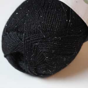 May include: A close-up of a skein of black yarn with silver flecks. The yarn is tightly wound and has a textured appearance. The label is partially visible. This yarn is suitable for knitting and crochet projects.