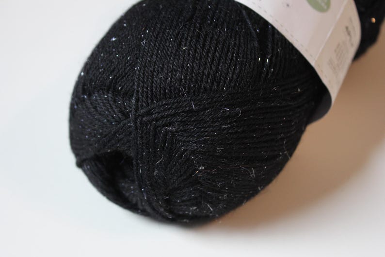 May include: A close-up of a skein of black yarn with silver flecks. The yarn is tightly wound and has a textured appearance. The label is partially visible. This yarn is suitable for knitting and crochet projects.