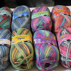 May include: Eight skeins of Opal yarn arranged in two rows of four. Each skein features a unique multi-colored variegated pattern and a paper label with the text 30 Jahre Opal. The yarn balls display various color combinations including blue, pink, green, and orange tones.