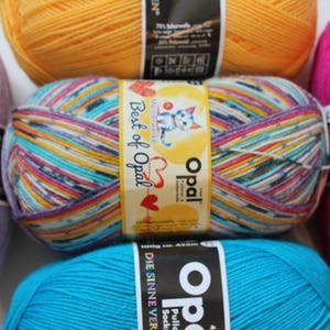 May include: Close-up of several colorful yarn skeins. The central skein features a label with the text "Best of Opal" and a cartoon cat. Other skeins are in shades of yellow, pink, blue, and purple. The yarn is likely for knitting or crochet projects.