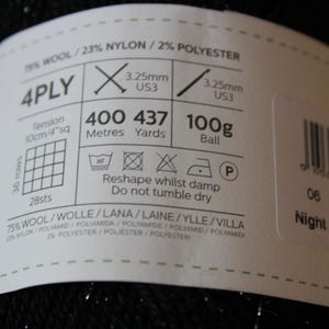May include: A label wrapped around a ball of black yarn. The label indicates the yarn is 4-ply, 75% wool, and 23% nylon. It includes knitting needle size, 400 meters of yarn, and care instructions. The text "Night Sky 1108" is also visible.