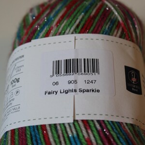 May include: A close-up of a ball of yarn with a label that reads "Fairy Lights Sparkie." The yarn is a mix of colors including red, green, blue, and silver. The label also includes a barcode and other text.