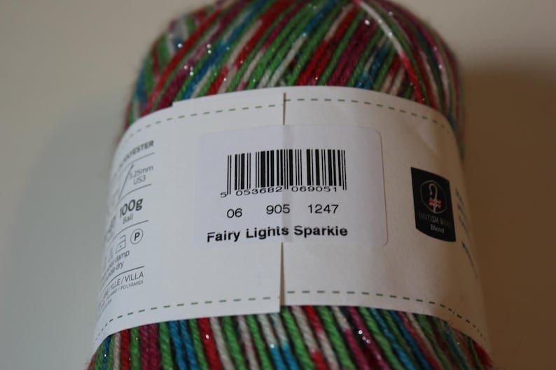 May include: A close-up of a ball of yarn with a label that reads "Fairy Lights Sparkie." The yarn is a mix of colors including red, green, blue, and silver. The label also includes a barcode and other text.