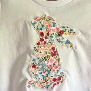 May include: White t-shirt with a floral bunny appliqué. The bunny is outlined in white stitching. The t-shirt has a brown tag with the text "Lily & Ro" on it.