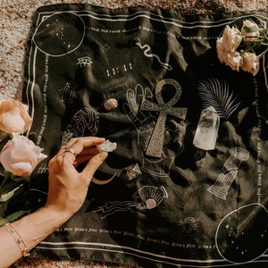 May include: A black cloth with white designs, including an ankh symbol, a snake, and a palm tree. There are several crystals and stones on the cloth, including a clear quartz crystal and a grey stone. The cloth is laid out on a sandy surface.