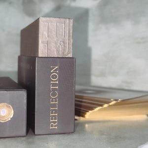 Op de afbeelding: Twee rechthoekige donkerbruine doosjes met gouden letters. E&eacute;n doosje staat rechtop met het woord REFLECTION verticaal in goud gedrukt. Het andere doosje ligt horizontaal met een rond gouden zegel op het uiteinde. Een stapel kaarten met gouden randen ligt ernaast.