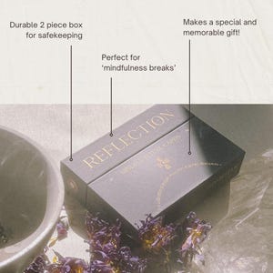 Op de afbeelding: Een zwarte rechthoekige tweedelige doos met de gouden tekst REFLECTION MINDFUL RITUAL CARDS. De doos bevat een klein gouden kruissymbool en de tekst a collection of simple mindful gentle disruptors. De doos staat in de buurt van een kom.