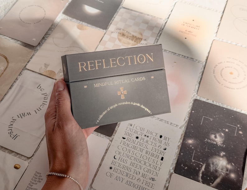 Op de afbeelding: Een donkergrijze rechthoekige doos met het label REFLECTION MINDFUL RITUAL CARDS in gouden letters en een klein gouden kruissymbool. De doos bevat de tekst a collection of simple reminders & gentle disruptions in een boog onder de titel.