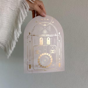 May include: A white card with a gold geometric design. The design features a circle with a series of smaller circles around it, as well as other geometric shapes.