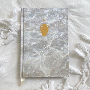 Puede incluir: Un diario con un patrón de mármol blanco con una luna creciente dorada y un diseño de urna dorada en la portada.