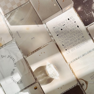 Op de afbeelding: Een verzameling rechthoekige orakelkaarten in beige en cr&egrave;metinten, voorzien van zwarte typografie en hemelse symbolen. E&eacute;n kaart bevat de tekst: I CALL BACK MY ENERGY FROM ALL TIMES, PLACES & REALITIES. Een helder, ruw kwartskristal ligt op een kaart.