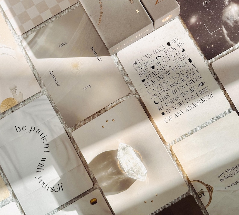 Op de afbeelding: Een verzameling rechthoekige orakelkaarten in beige en cr&egrave;metinten, voorzien van zwarte typografie en hemelse symbolen. E&eacute;n kaart bevat de tekst: I CALL BACK MY ENERGY FROM ALL TIMES, PLACES & REALITIES. Een helder, ruw kwartskristal ligt op een kaart.