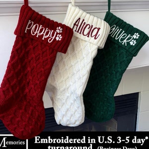 May include: Three personalized Christmas stockings in red, white, and green, each with a name embroidered on the cuff. The red stocking says "Poppy", the white says "Alicia", and the green says "Oliver". Each stocking has a paw print design.