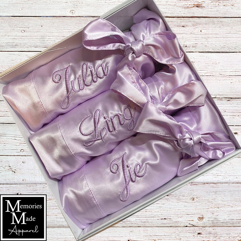 May include: A set of three lilac satin robes, each embroidered with a name: "Julia", "Ling", and "Jie", presented in a white box. Each robe features a matching satin bow. The box also displays the "Memories Made Apparel" logo.