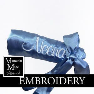 May include: A light blue satin sash embroidered with the name "Neena" in white script. The sash is tied in a bow and features the text "EMBROIDERY" in a black banner. The logo "Memories Made Apparel" is in a white square.