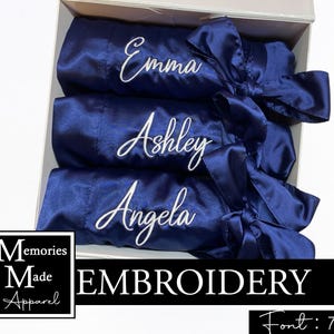 May include: Three navy blue satin robes, each embroidered with a name in white: Emma, Ashley, and Angela, are presented in a white box. Each robe features a matching bow. The image also displays the text "Memories Made Apparel" and "EMBROIDERY".