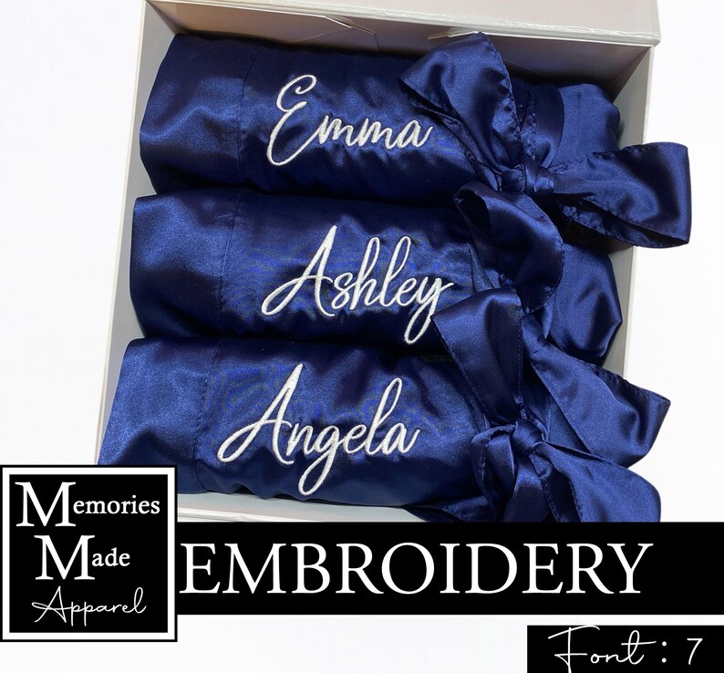 May include: Three navy blue satin robes, each embroidered with a name in white: Emma, Ashley, and Angela, are presented in a white box. Each robe features a matching bow. The image also displays the text "Memories Made Apparel" and "EMBROIDERY".