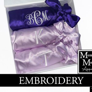 May include: A white gift box contains three satin robes in purple and lavender. Each robe is embroidered with a single initial or monogram in white thread. The robes are tied with matching satin bows. The box has the text "EMBROIDERY" and a logo.