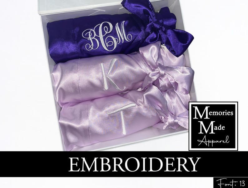 May include: A white gift box contains three satin robes in purple and lavender. Each robe is embroidered with a single initial or monogram in white thread. The robes are tied with matching satin bows. The box has the text "EMBROIDERY" and a logo.