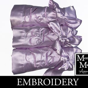 May include: Three lilac satin robes, each embroidered with initials and tied with matching bows. The robes are stacked, displaying the initials "KA", "ER", and "VB". The image also features the word "EMBROIDERY" and the "Memories Made Apparel" logo.