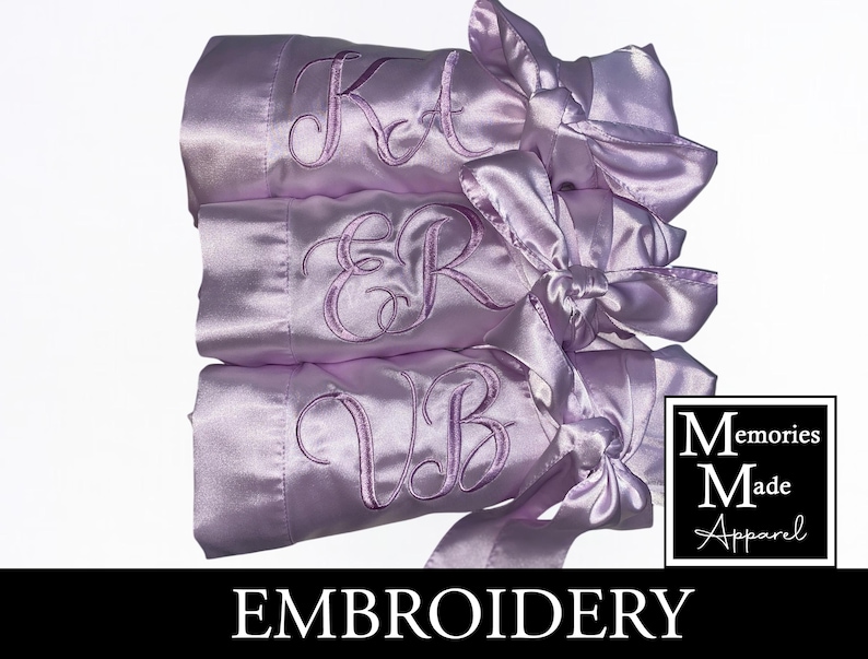 May include: Three lilac satin robes, each embroidered with initials and tied with matching bows. The robes are stacked, displaying the initials "KA", "ER", and "VB". The image also features the word "EMBROIDERY" and the "Memories Made Apparel" logo.