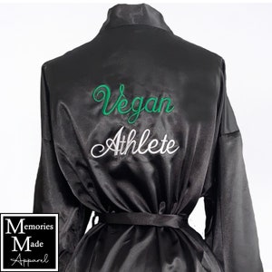 May include: Black satin robe with a matching belt. The back of the robe features the embroidered text "Vegan Athlete" in green and white script. The robe is displayed on a mannequin against a white background.