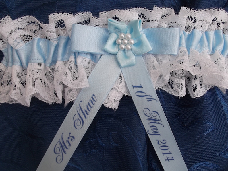 K&ouml;nnte beinhalten: Ein blaues und wei&szlig;es Strumpfband mit einem blauen Satinband und einer wei&szlig;en Spitzenborte. Das Band ist mit dem Text "Mrs. Shaw" und "10th May 2014" personalisiert.