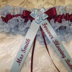 May include: A blue and burgundy garter with a blue satin bow and pearl embellishment. The garter has two blue satin ribbons with the text "Forever Blowing Bubbles" and "Mrs. Smith" printed in burgundy.