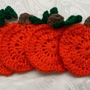 May include: A set of four circular orange crochet coasters, each featuring a brown stem and green leaves at the top. The coasters are crafted with a textured yarn pattern, creating a pumpkin-like appearance. The items are arranged in a horizontal row.