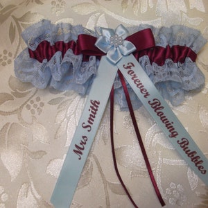 May include: A light blue garter with a burgundy satin bow and a light blue flower with a pearl center. The garter has two light blue satin ribbons attached, one with the text "Mrs. Smith" and the other with the text "Forever Blowing Bubbles".