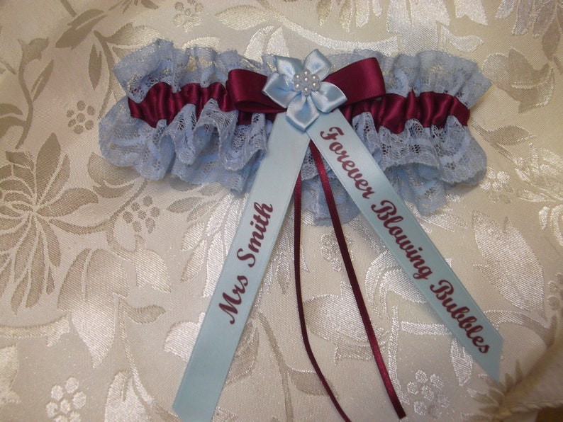 May include: A light blue garter with a burgundy satin bow and a light blue flower with a pearl center. The garter has two light blue satin ribbons attached, one with the text "Mrs. Smith" and the other with the text "Forever Blowing Bubbles".