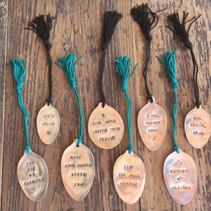 May include: A collection of handmade spoon bookmarks with stamped text and colorful tassels. The spoons have phrases like "I like books better than people" and "I'd rather be reading". The tassels are black and teal.