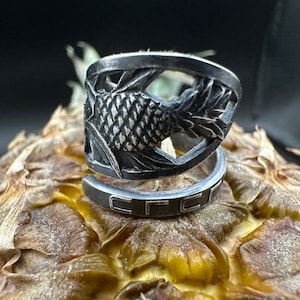 May include: A silver-colored ring with a pineapple design. The ring features a detailed carving of a pineapple and leaves. The ring is set on a pineapple, with a dark background.