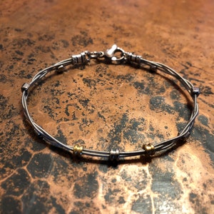 May include: A silver bracelet made from twisted metal strands, featuring small silver and gold-colored beads. The bracelet has a clasp closure and is displayed on a textured, copper-colored surface. Jewelry.