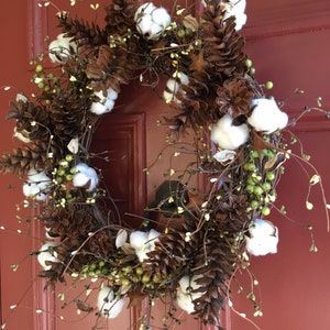 May include: A brown and white wreath with pine cones and cotton balls. The wreath is hanging on a red door.