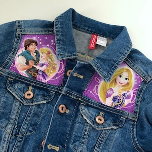 Puede incluir: Chaqueta vaquera azul con dos parches con personajes de cuento de hadas. Un parche muestra una pareja y el otro una princesa. La chaqueta tiene botones color cobre y un cuello clásico.