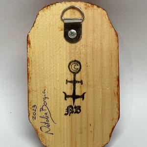 K&ouml;nnte beinhalten: Die R&uuml;ckseite eines Holzgem&auml;ldes mit einem Metall-Aufh&auml;nger. Das Gem&auml;lde ist signiert mit "Natalia Borgia" und datiert mit "2023". Es gibt ein Symbol mit einem Kreuz und den Buchstaben "J.P.B."