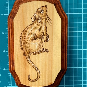 K&ouml;nnte beinhalten: Eine Holzplakette mit braunem Rahmen zeigt ein eingebranntes Bild einer Ratte, die nach oben schaut. Die Ratte ist mit Linien und Schattierungen detailliert.