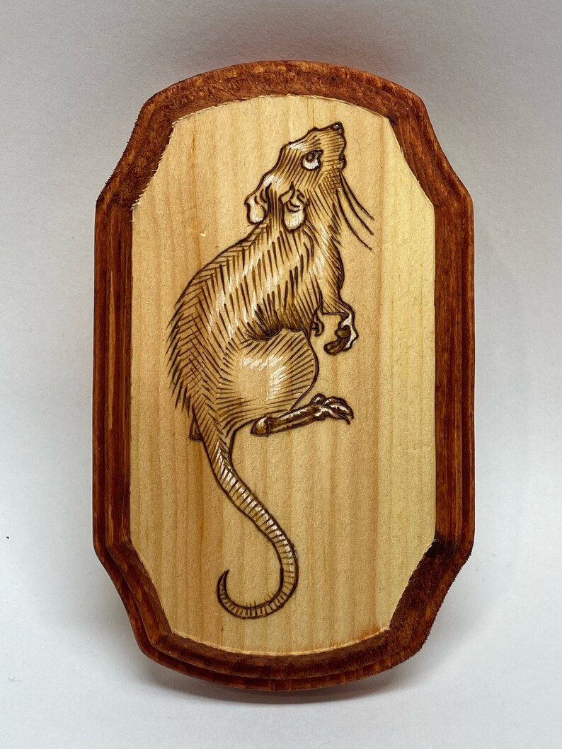K&ouml;nnte beinhalten: Ein Holzbrandbild einer Ratte auf einer Holztafel mit rotem Rand.