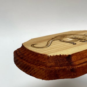 K&ouml;nnte beinhalten: Eine Holzplakette mit einem lasergravierten Bild eines Dinosauriers. Die Plakette hat einen dunkelbraunen Rand und eine hellbraune Mitte.