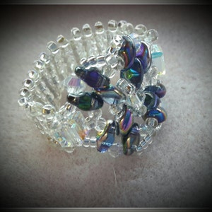 May include: A beaded ring with a flower design made of iridescent blue and purple beads. The ring is made of clear beads and is adjustable.