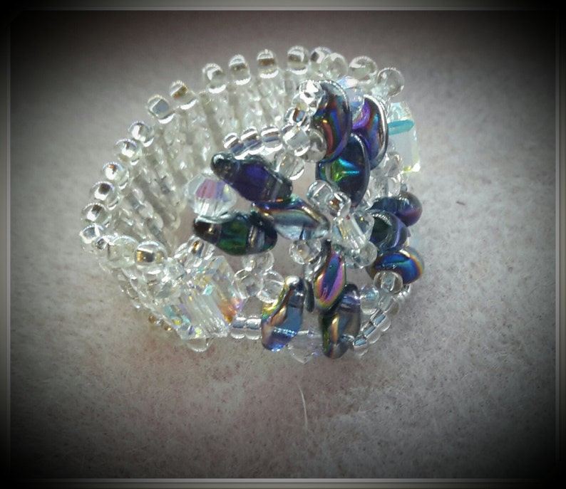 May include: A beaded ring with a flower design made of iridescent blue and purple beads. The ring is made of clear beads and is adjustable.