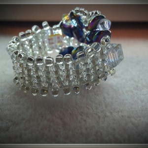 May include: A silver ring with a cluster of iridescent blue and clear glass beads. The ring is made of small, square beads that are arranged in a pattern.