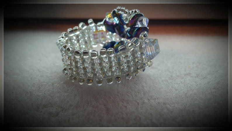 May include: A silver ring with a cluster of iridescent blue and clear glass beads. The ring is made of small, square beads that are arranged in a pattern.