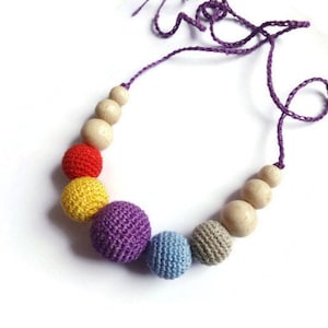 May include: A colorful necklace featuring a mix of wooden and crocheted beads. The necklace has a purple cord and includes red, yellow, purple, blue, and gray crocheted beads, along with natural wooden beads. The beads are strung together to create a unique accessory.