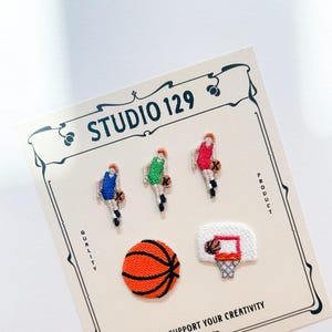 Puede incluir: Cinco parches bordados para planchar que representan jugadores de baloncesto, una pelota de baloncesto y una canasta de baloncesto. Los parches están en una tarjeta blanca con el texto "Studio 129" y "Apoyamos tu creatividad".